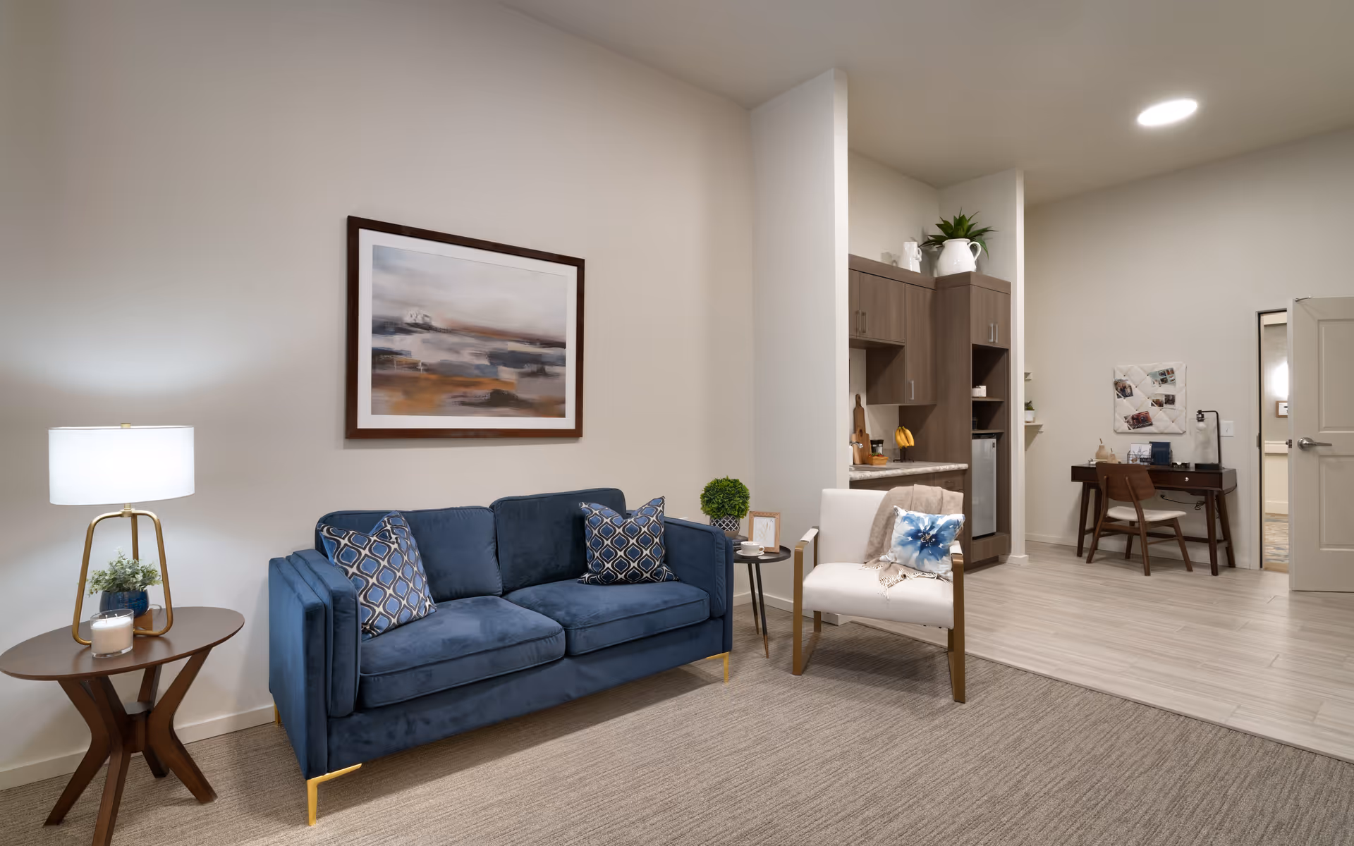 Furnished living area with a blue sofa, armchair, side tables and lamp, wall art, and a kitchenette and desk in the background.