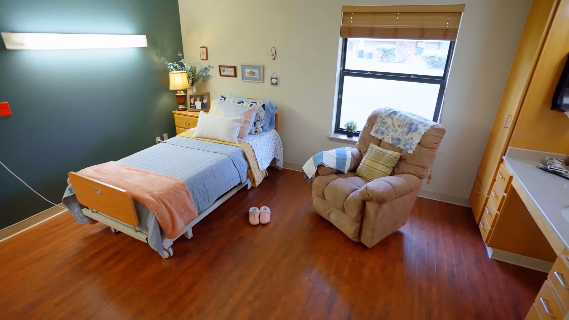 A cozy single bedroom in a care facility with a bed made up with blue and white bedding, a wooden nightstand with a lamp and framed photos, a comfortable recliner chair with a throw blanket and pillow, a window with blinds, and wooden flooring.