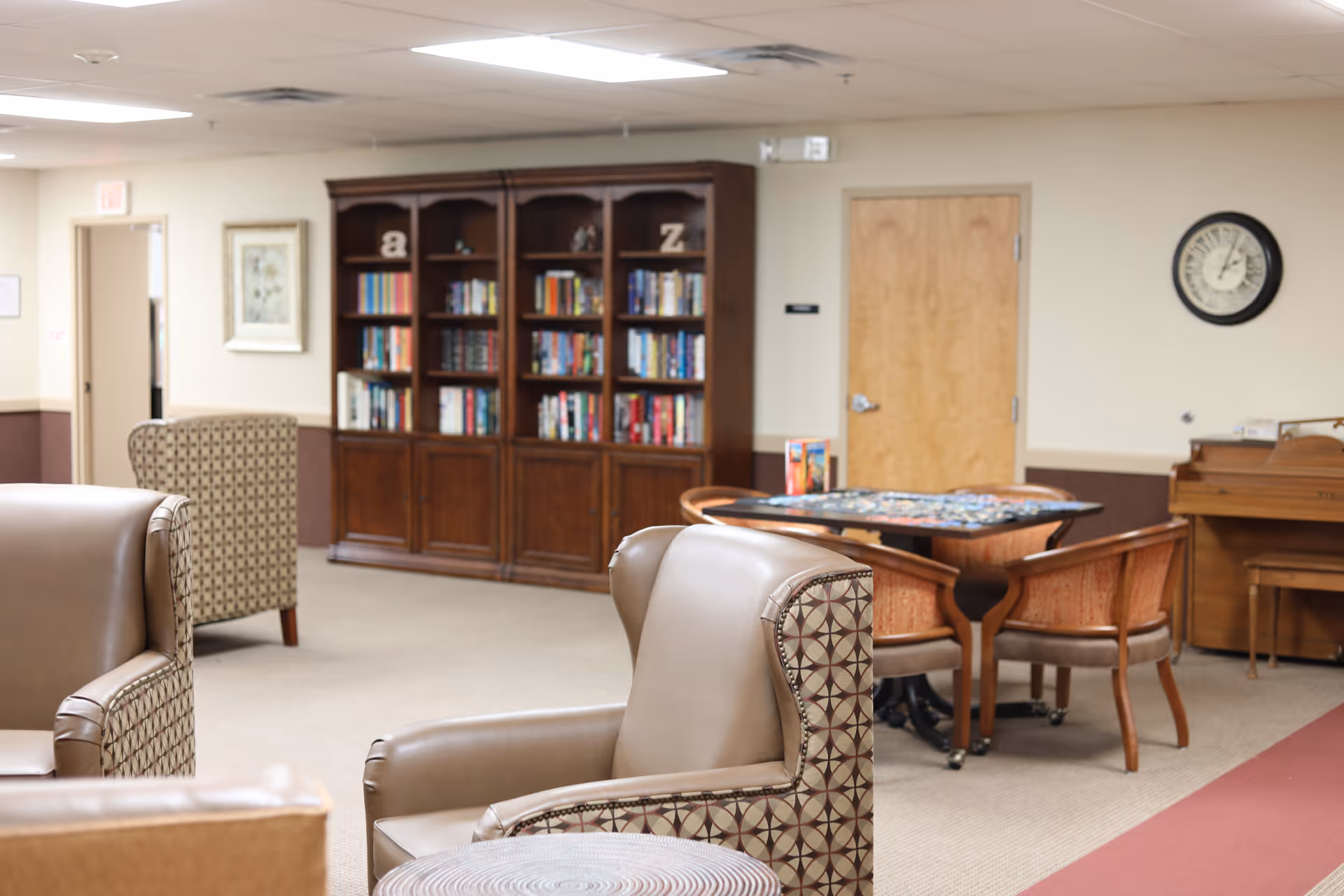 A cozy common area in a senior living facility featuring comfortable armchairs with patterned upholstery, a wooden bookshelf filled with books, a round table with four chairs, a puzzle in progress on the table, a piano against the wall, a clock showing the time, and a wooden door.