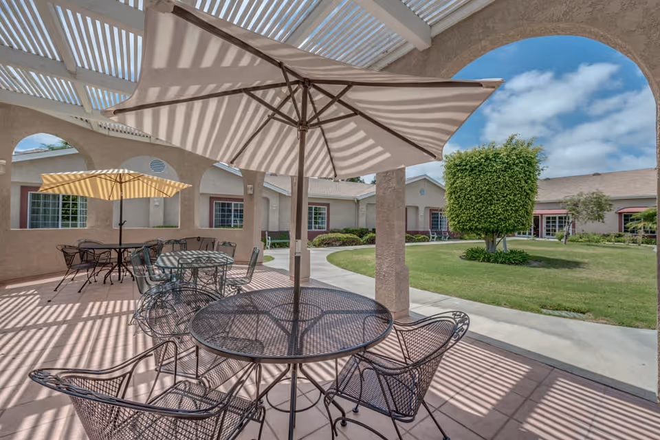 Outdoor patio area at Ivy Terrace at Garden Grove Memory Care featuring metal tables and chairs with large umbrellas providing shade. The patio is covered with a pergola casting striped shadows. In the background, there is a well-maintained lawn, a trimmed tree, and the exterior of the facility building under a partly cloudy sky.
