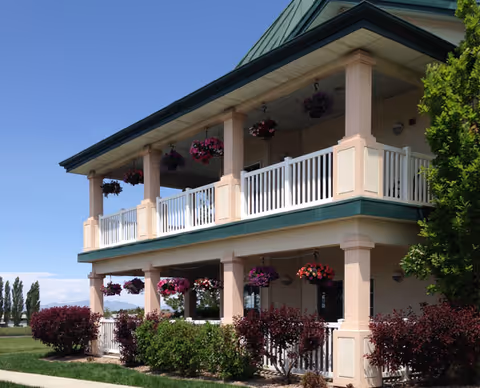 Two-story pale pink building with covered balconies, white railings, hanging flower baskets and landscaped shrubs under a clear blue sky.