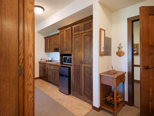 Small interior kitchenette with dark wood cabinets, a microwave and mini-fridge, and a side table with decorative items near a doorway.