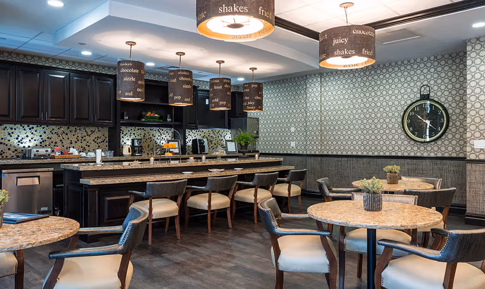 A modern dining area with round tables and cushioned chairs, a long counter with bar stools, dark wood cabinetry, and pendant lights hanging from the ceiling. The walls have patterned wallpaper, and there is a large round clock on one wall.
