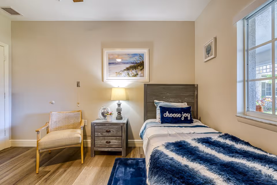 A cozy bedroom with a single bed featuring a blue and white striped blanket and a pillow with the words 'choose joy'. Next to the bed is a wooden nightstand with a lamp, a small fan, and a small plant. A wooden chair with a woven backrest is placed beside the nightstand. The room has beige walls, a window showing an outdoor view, and framed artwork on the walls.