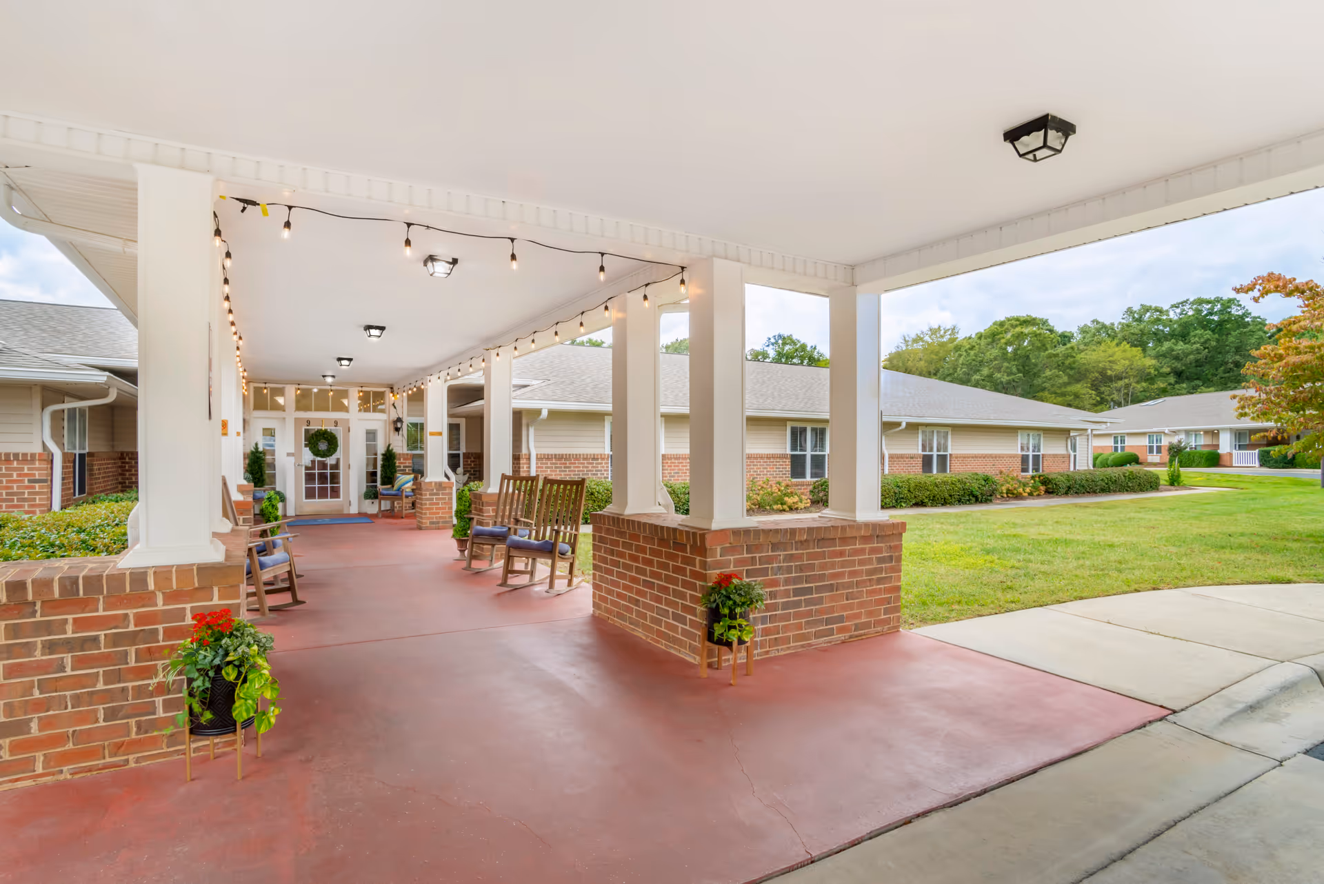 Covered front entrance/portico with brick columns, rocking chairs, string lights, potted plants, and a view of the surrounding lawn and building.