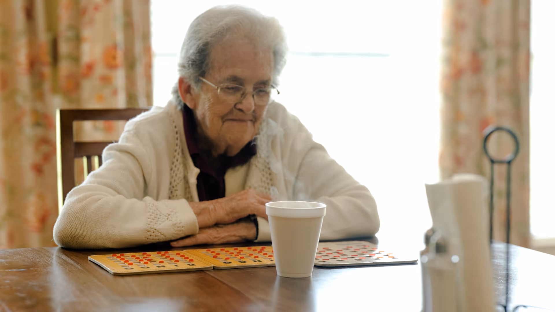 An elderly woman with glasses and white hair sits at a wooden table playing bingo. She is wearing a white cardigan and looking down at her bingo cards with a slight smile. A white disposable cup is on the table in front of her, and floral curtains are visible in the background.