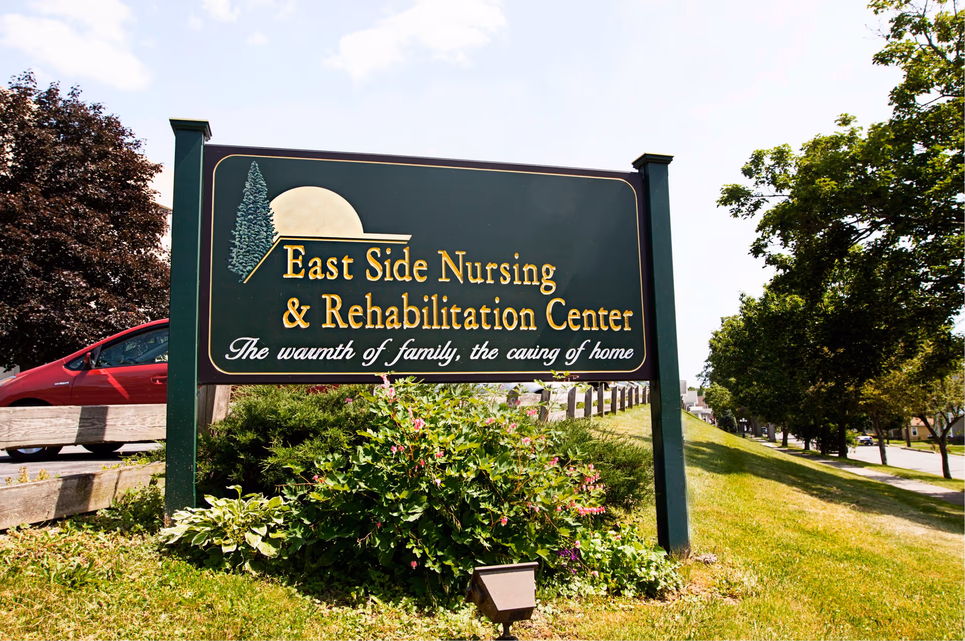 Outdoor sign for East Side Nursing & Rehabilitation Center with the tagline 'The warmth of family, the caring of home', surrounded by greenery and trees along a sidewalk.