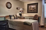 A cozy bedroom in a senior living facility featuring a single bed with a beige bedspread and pillows, a wooden nightstand with a lamp and a vase of flowers, a round wicker chair, a wall clock, and a framed landscape painting. The room has soft lighting and a window with curtains.