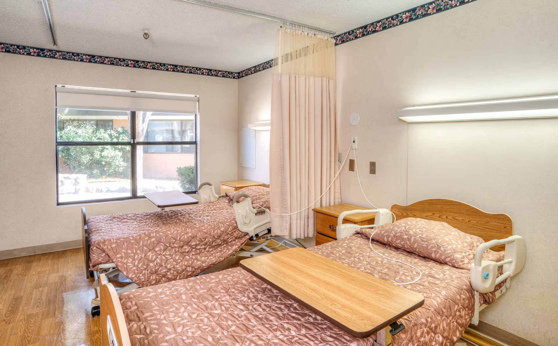 A room with two hospital-style beds, each with a brown patterned bedspread and a wooden headboard. Both beds have adjustable side rails and overbed tables. There is a window with a view of greenery outside, a curtain divider between the beds, and a floral border near the ceiling. The room has wooden flooring and soft lighting above each bed.