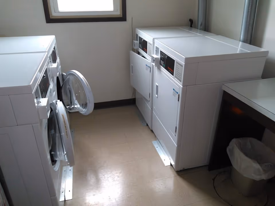 Laundry room with two front-loading washing machines on the left, one with its door open, and two front-loading dryers on the right. There is a window above the washing machines and a trash can under a counter on the right side.