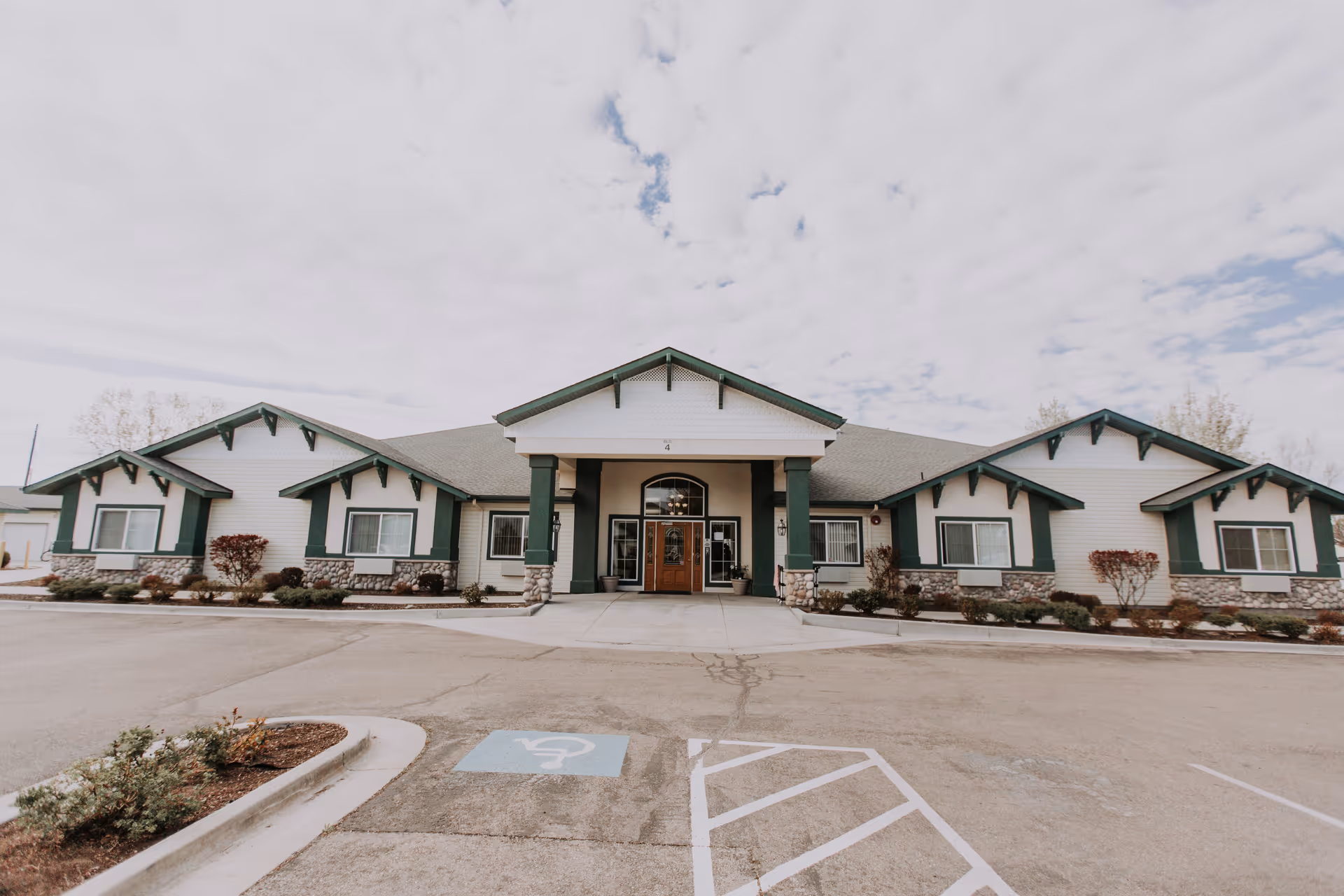 Front exterior view of Swan Falls Assisted Living facility showing a single-story building with green and white trim, stone accents, a covered entrance with double wooden doors, and a parking lot with a handicap parking space in front.