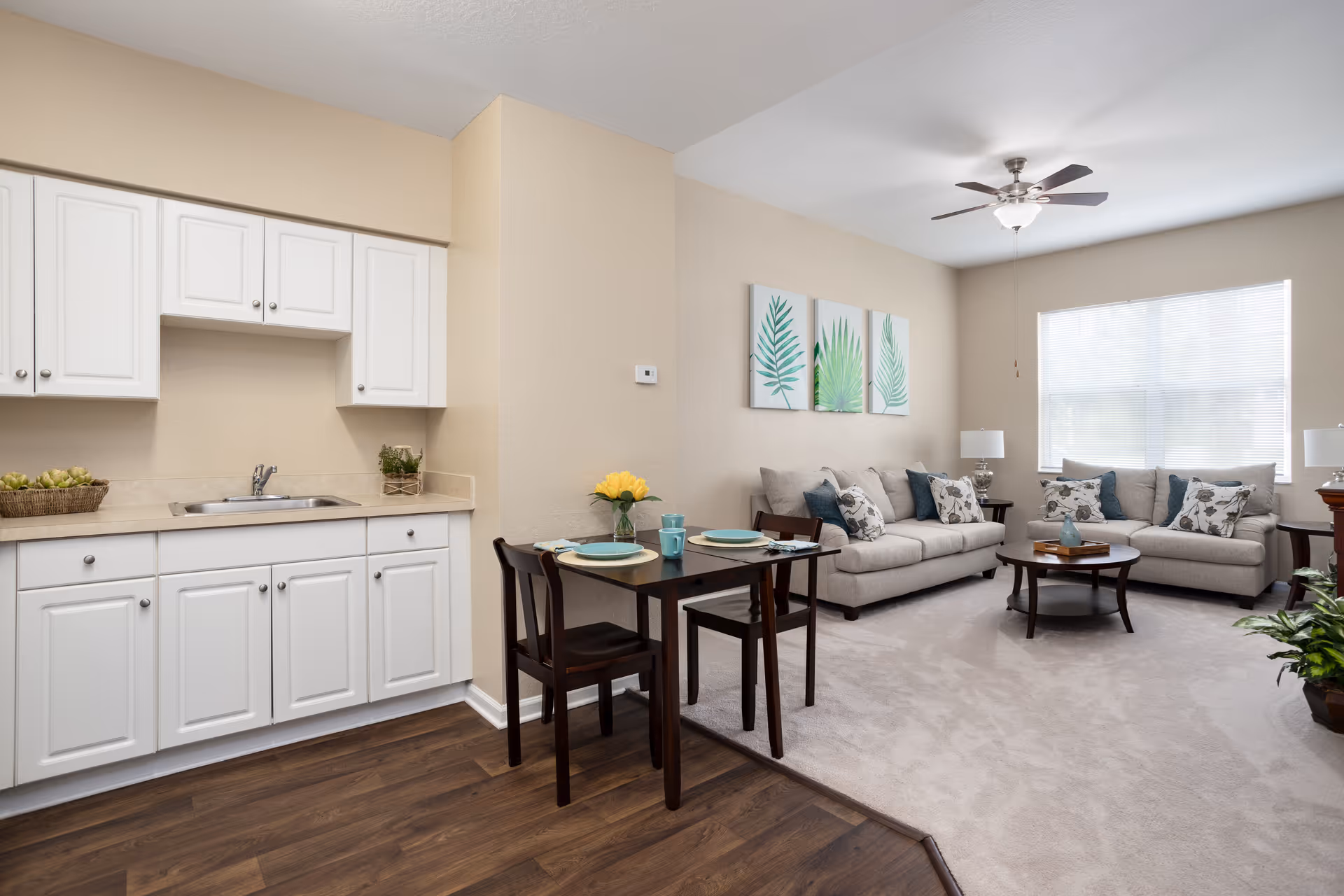 A bright and clean living space in a senior living facility featuring a small kitchen area with white cabinets and a sink, a dark wooden dining table set for two with plates and a vase of yellow flowers, and a cozy living room with two beige sofas adorned with patterned cushions, a round wooden coffee table, two table lamps, and three framed botanical prints on the wall. A ceiling fan with a light fixture is mounted on the ceiling, and a large window with blinds allows natural light to fill the room.