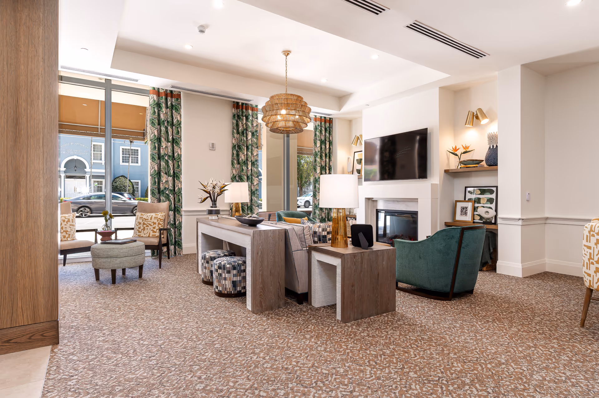 Bright furnished common living room with seating, a TV above a fireplace, decorative lamps, and large windows facing the street.
