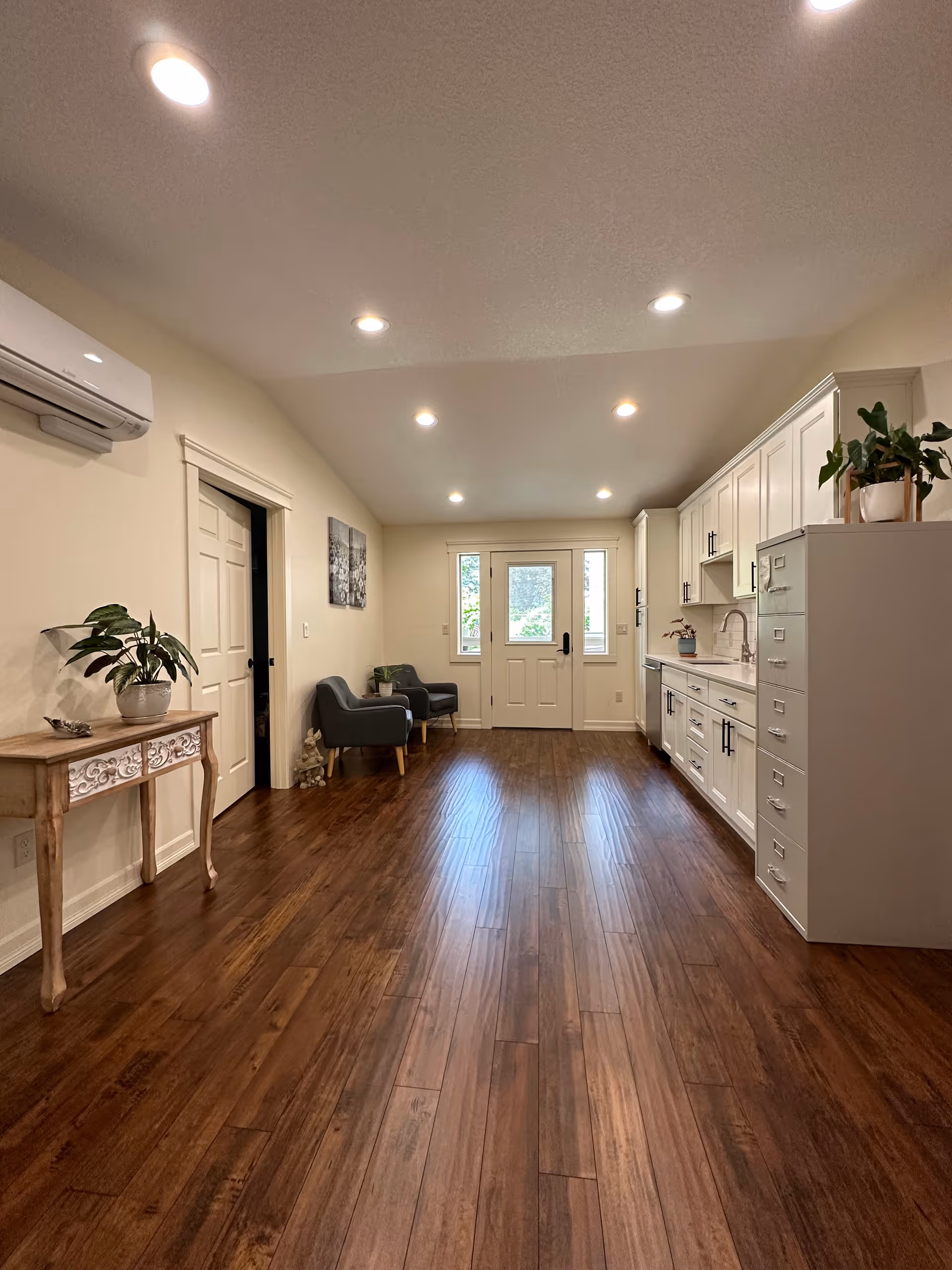 Interior view of a room with wooden flooring, white cabinets and countertop on the right side, two gray armchairs and a small table near a door with windows at the far end, a decorative wooden table with a potted plant on the left, and recessed ceiling lights.