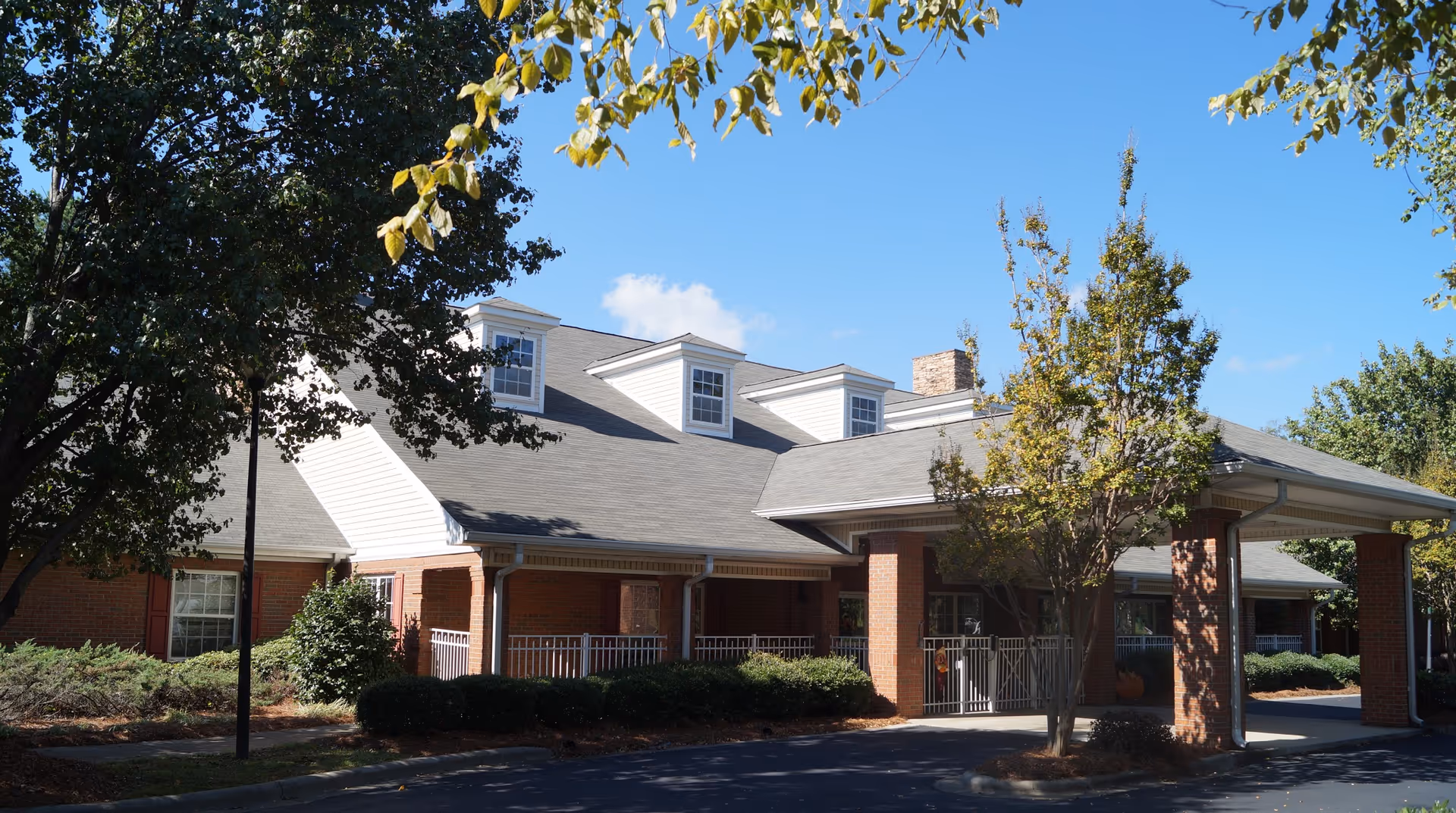 Front entrance of a brick senior living facility with a covered porte-cochere, dormer windows, and landscaping under a blue sky.