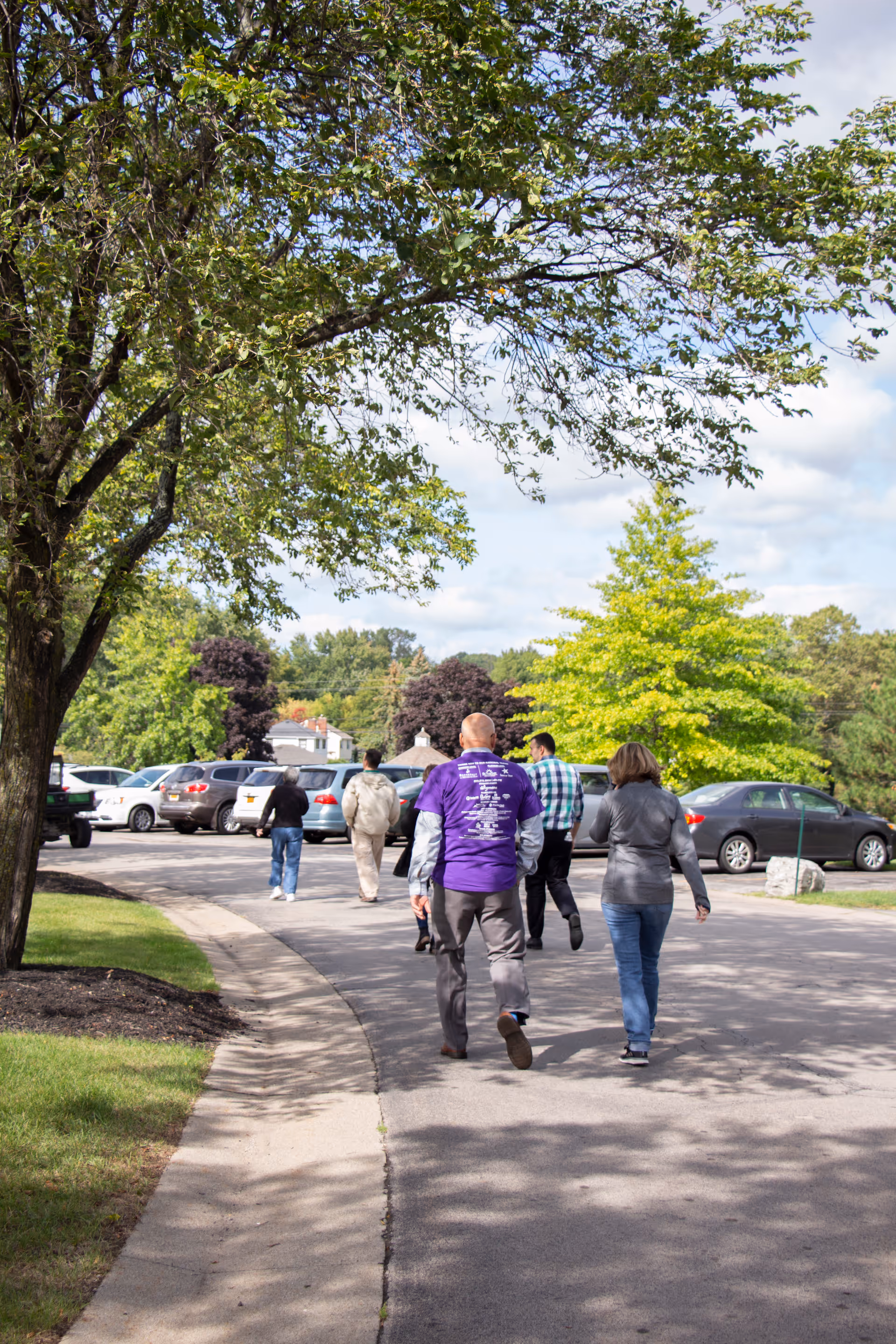 A group of people walking on a paved pathway in a parking lot surrounded by trees and parked cars on a sunny day.