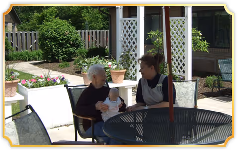 Two people sit at a round outdoor patio table surrounded by potted flowers, lattice panels, and garden landscaping.