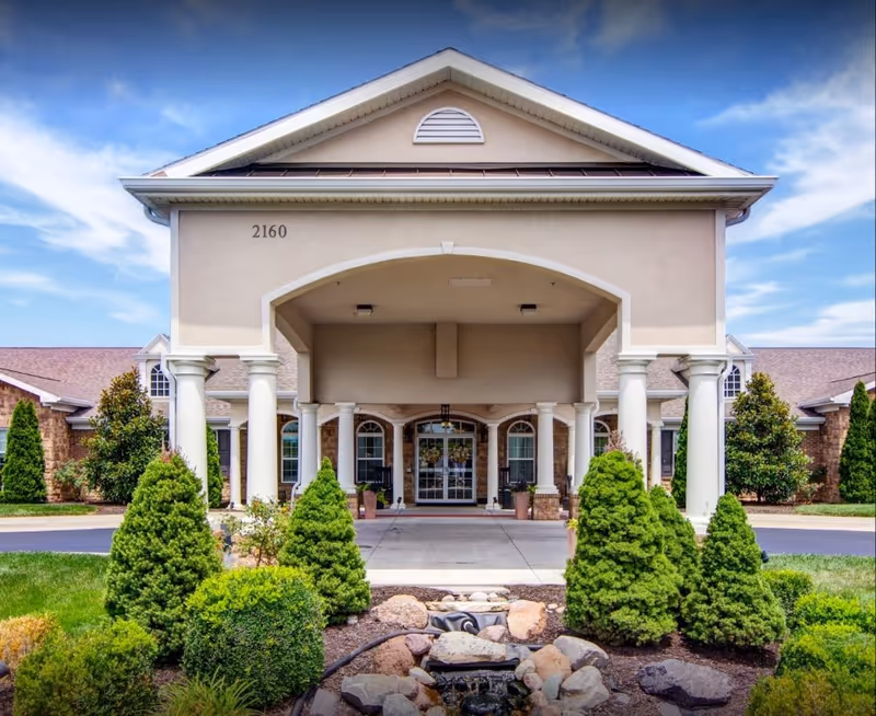 Front exterior view of Addington Place of Lees Summit building with a covered entrance supported by white columns, surrounded by neatly trimmed bushes and landscaping under a blue sky with some clouds.