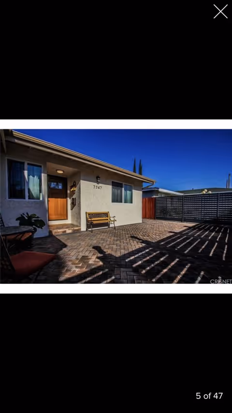 Outdoor patio area of a residence with a paved brick floor, a wooden bench against the wall, a wooden door, two windows, and a privacy fence. The house number 7747 is visible on the wall next to the door. The sky is clear and blue.