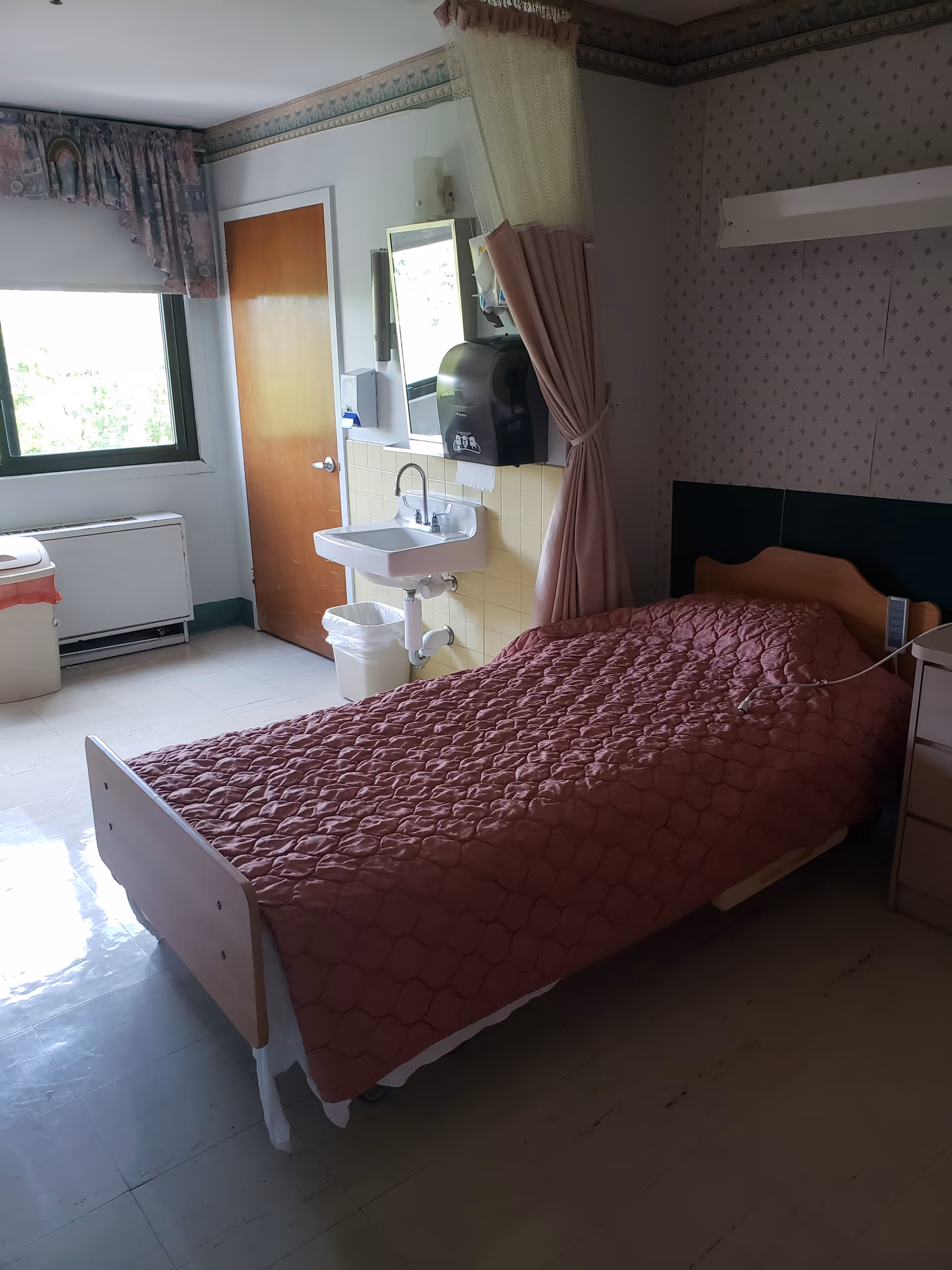 A small room with a single bed covered with a pink quilted blanket. Next to the bed is a nightstand. There is a sink with a mirror above it and a paper towel dispenser mounted on the wall. A curtain is partially drawn around the bed area. A window with floral curtains and a wooden door are visible in the background. The floor is tiled and there is a trash bin near the sink.
