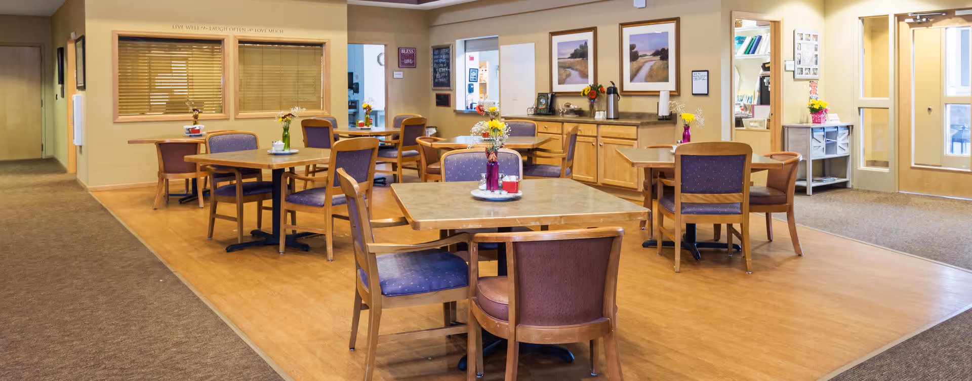 A bright and welcoming dining area in an assisted living facility with several square wooden tables and chairs with purple cushions. Each table has a small vase with flowers. The room features light wood flooring, beige walls, framed landscape pictures, and a sideboard with coffee and tea supplies. There is a doorway leading to another room and a glass door entrance on the right side.