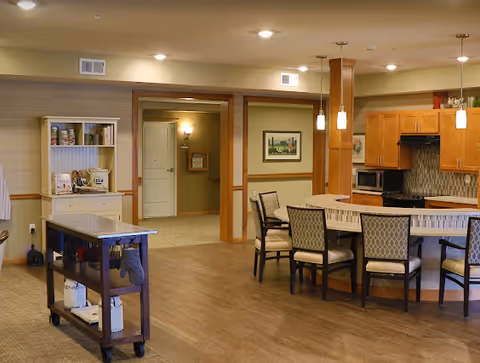 Interior view of a senior living facility kitchen and dining area with wooden floors, a curved counter with chairs, wooden cabinets, a microwave, and pendant lights. There is a small cart with kitchen items and a white shelving unit with canned goods. The background shows a hallway with a door and framed artwork on the walls.