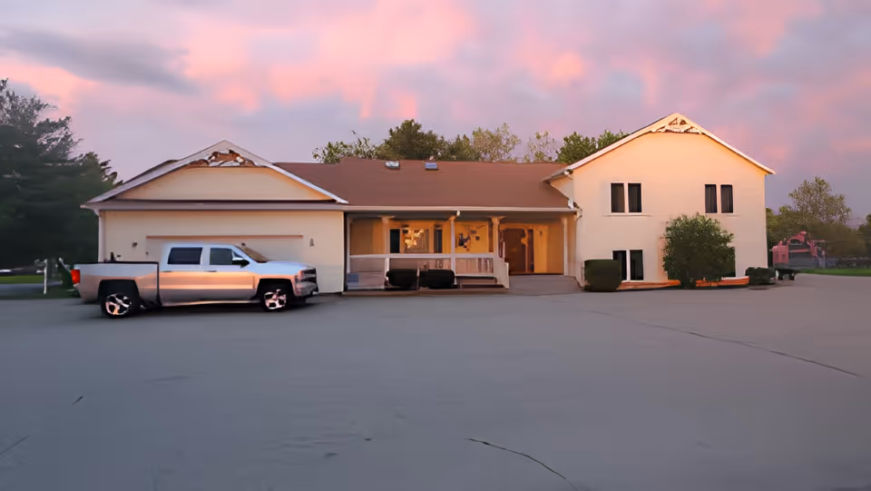 Exterior view of a single-story and two-story building with a brown roof and light-colored walls during sunset. A silver pickup truck is parked in front of the building on a large paved area. Trees and greenery surround the building under a pink and purple sky.
