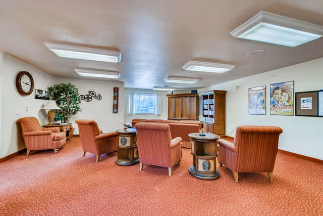 A cozy common area with six orange upholstered armchairs arranged around three round wooden side tables on a red carpeted floor. The room has white walls decorated with framed posters and a large clock. There is a wooden cabinet with shelves holding DVDs or books, a potted plant, and ceiling lights providing bright illumination.