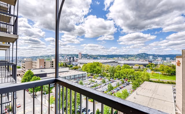 View from a balcony overlooking a parking lot, green trees, and a cityscape with buildings under a partly cloudy sky.