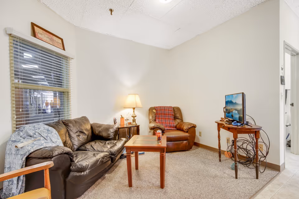 Small living room with a leather sofa, recliner, coffee table, lamp, and a TV on a wooden side table.