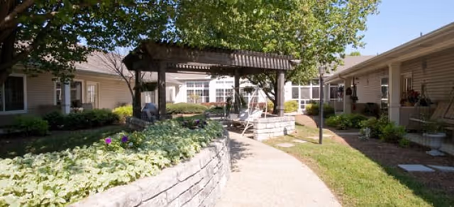 Outdoor courtyard area at Brookdale Gallatin featuring a curved concrete pathway, raised stone flower beds with green plants and purple flowers, a wooden pergola with seating underneath, and surrounding single-story buildings with beige siding and windows. Trees provide shade and the sky is clear and blue.