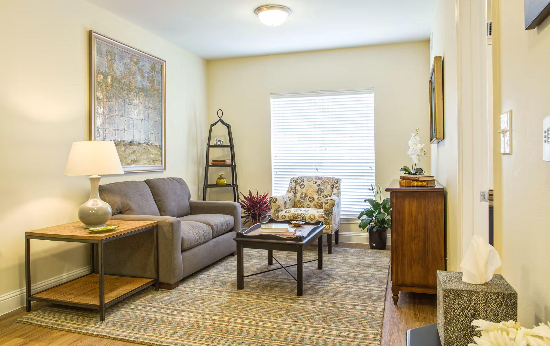 A cozy living room with a gray sofa, a patterned armchair, a wooden coffee table, and a side table with a lamp. The room has a large window with blinds, a tall black shelf with decorative items, a wooden cabinet with a plant and books, and a beige area rug on a wooden floor.