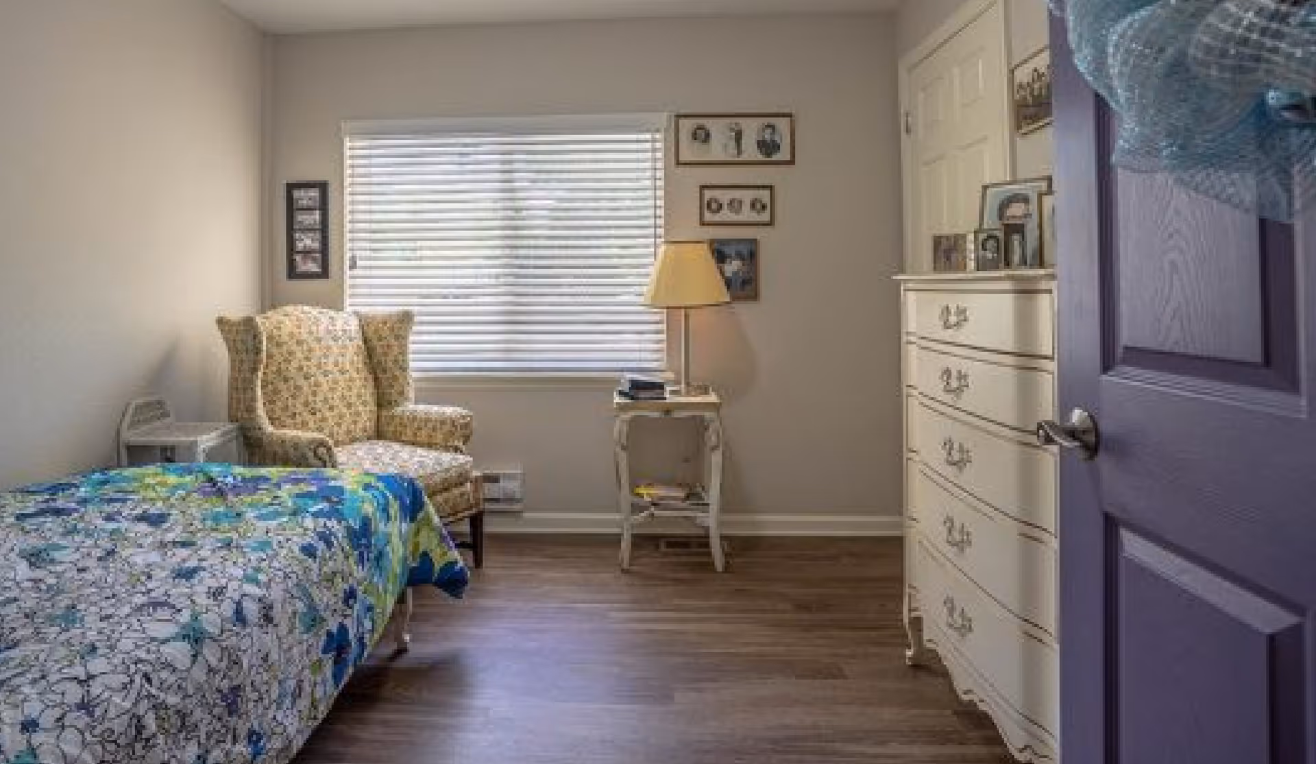 Small furnished bedroom with a floral bedspread, upholstered armchair, nightstand and dresser seen through a purple door.