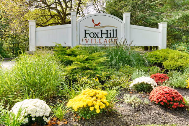 A white wooden sign with the text 'Fox Hill Village' surrounded by green shrubs and colorful flowers including yellow, white, and red blooms, with trees in the background.
