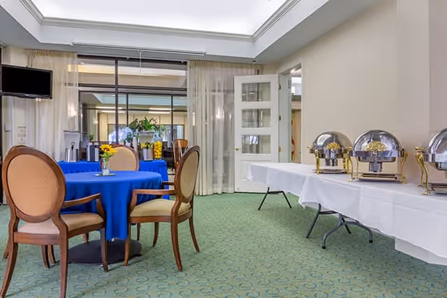 Dining room with round tables covered in blue tablecloths, wooden chairs, and a buffet table with chafing dishes.
