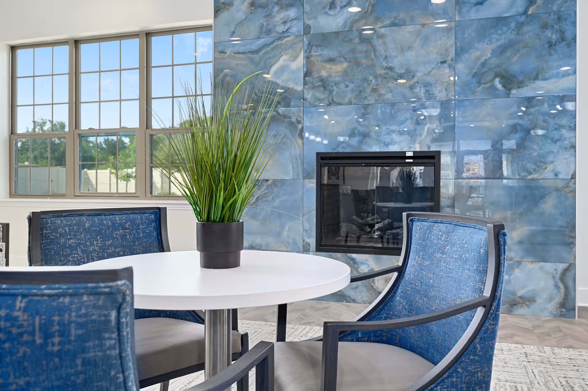 A cozy seating area with a round white table surrounded by blue upholstered chairs. A potted green plant is placed on the table. Behind the seating area is a modern fireplace set in a wall with blue marble-like tiles. Large windows let in natural light and show greenery outside.