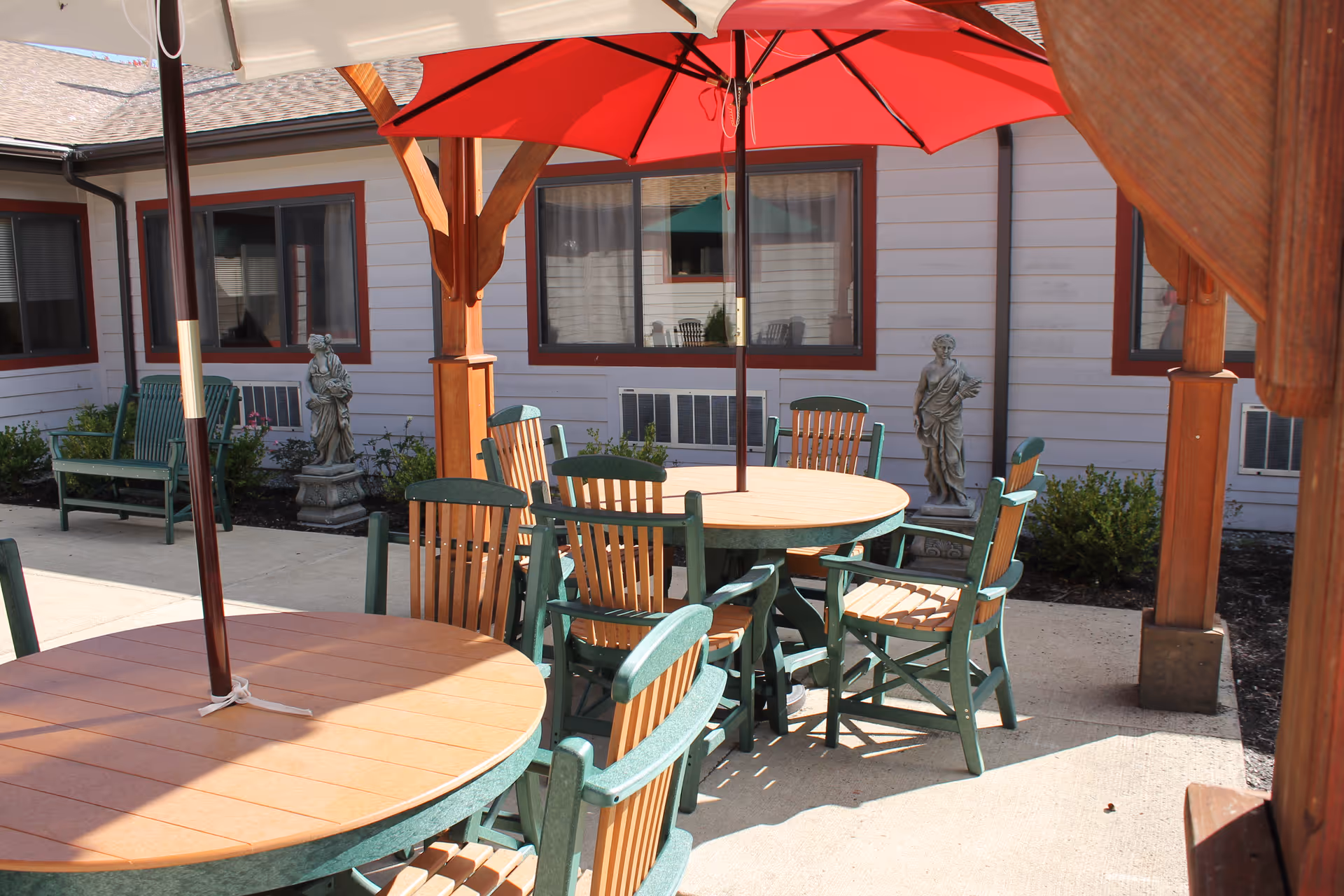 Outdoor courtyard with round wooden tables and green chairs under red umbrellas beside a building.