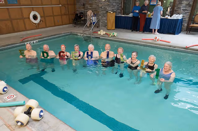 A group of elderly people standing in an indoor swimming pool holding up letters that spell 'EDGEHILL'. In the background, three people stand near a table with refreshments. The pool area has stone walls and pool equipment on the side.
