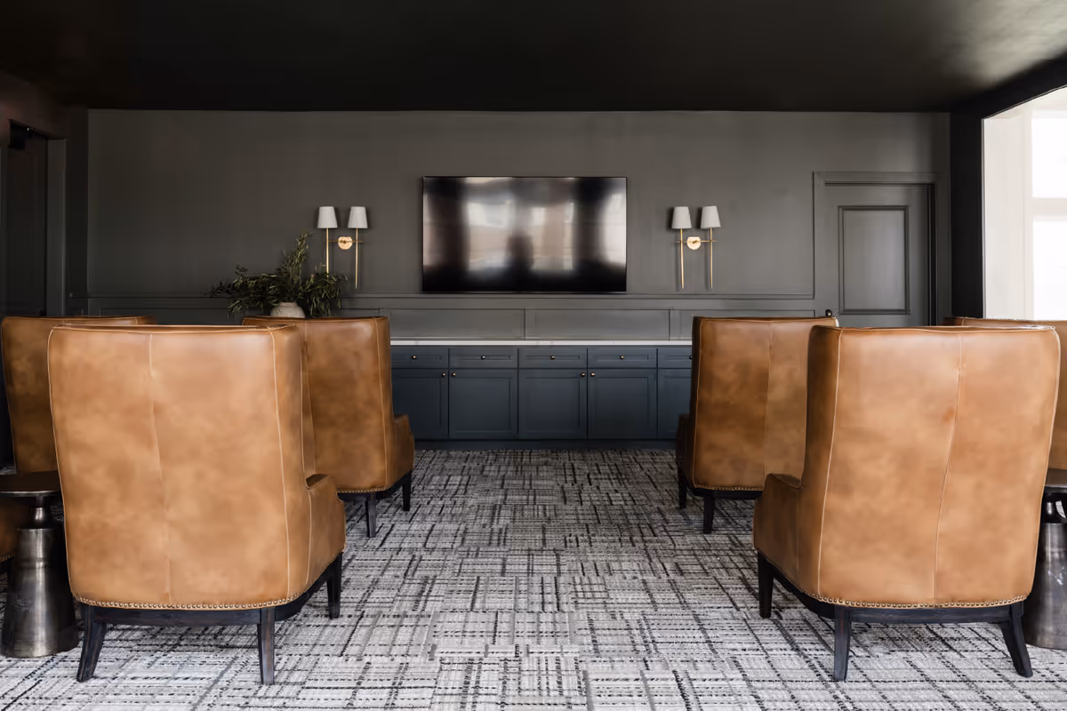 A modern lounge area with six brown leather armchairs arranged in two rows facing a large flat-screen TV mounted on a dark gray wall. Below the TV is a long blue cabinet with multiple drawers and doors. The room has a patterned carpet and two wall sconces with white lampshades flanking the TV. A potted plant is placed on the cabinet to the left.