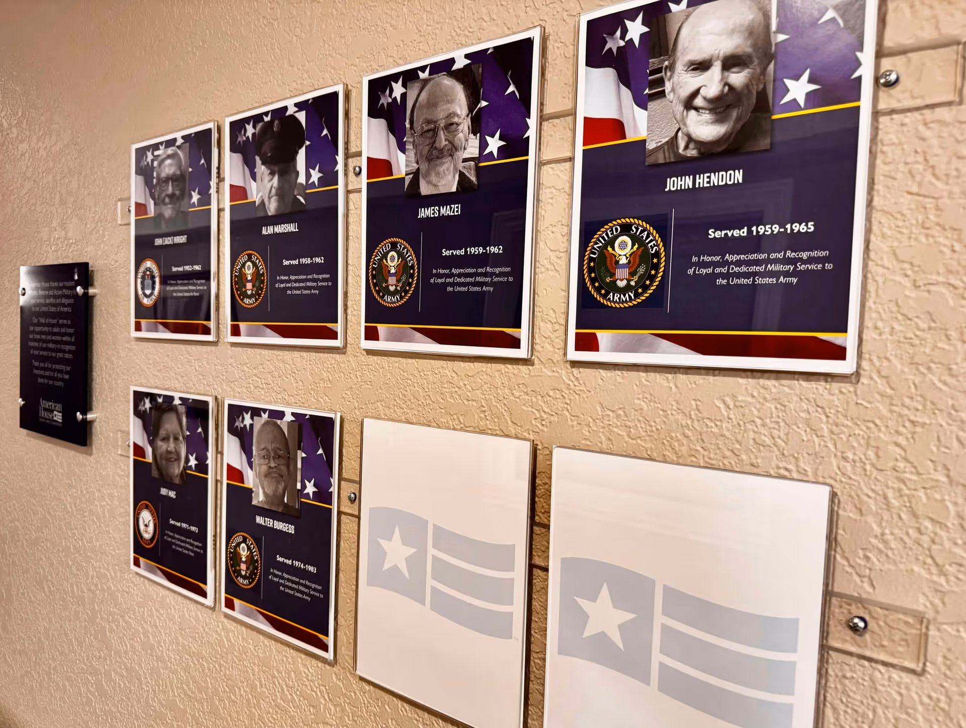 Wall display featuring framed photos and information plaques honoring military veterans with their names, service years, and United States Army emblems, mounted on a textured beige wall.