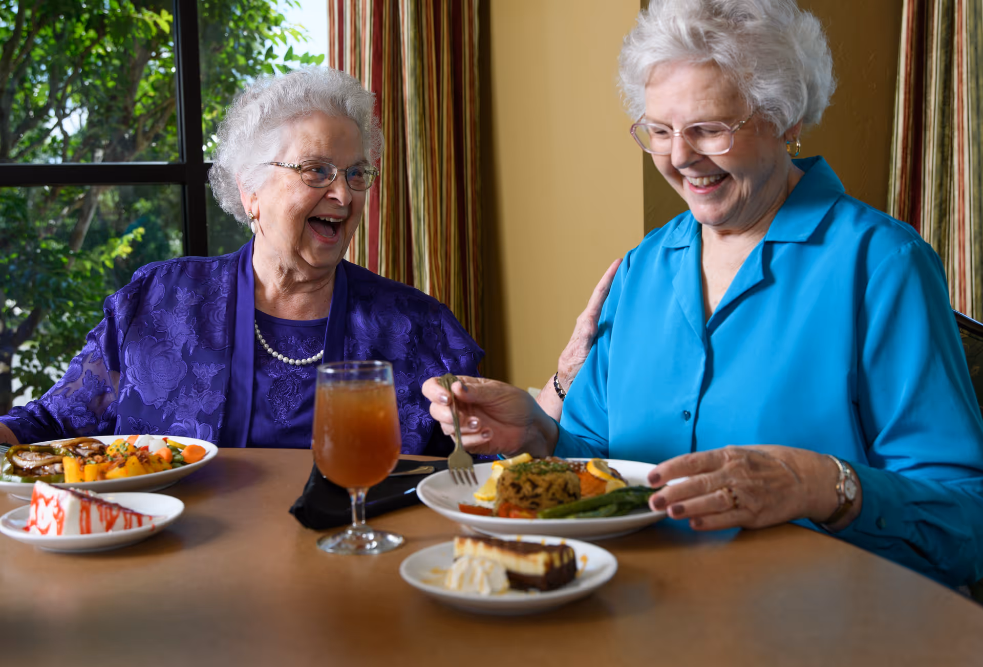 Two elderly women laughing and enjoying a meal together at a dining table in a bright room.