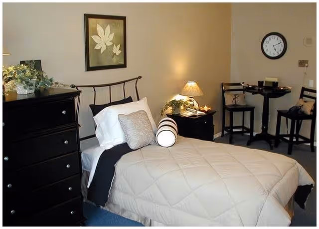 A neatly made single bed with a beige quilt in a furnished bedroom featuring a dark dresser, nightstand with a lamp, wall art, and a small table with two chairs.