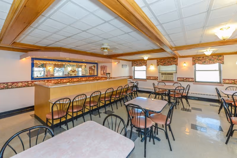 A dining room with multiple tables and chairs arranged neatly. There is a long counter with bar stools along one side. The room has a tiled ceiling with wooden beams, floral wallpaper border, and windows with floral valances. An air conditioning unit is visible in one window.