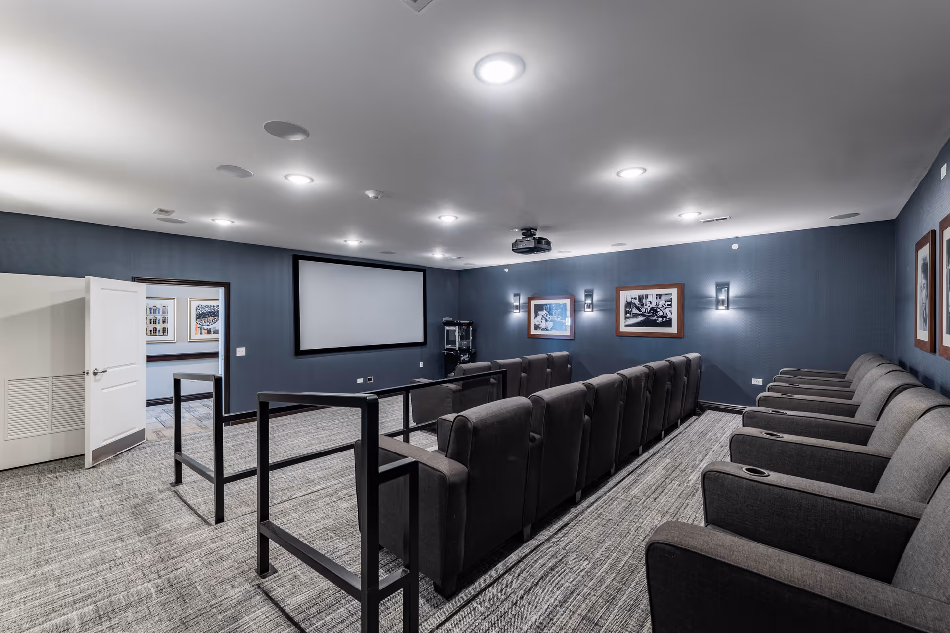A senior living facility movie theater room with two rows of gray upholstered reclining chairs facing a large blank projection screen on a blue wall. The room has a carpeted floor, recessed ceiling lights, framed black and white photos on the walls, and a popcorn machine in the corner near an open door.