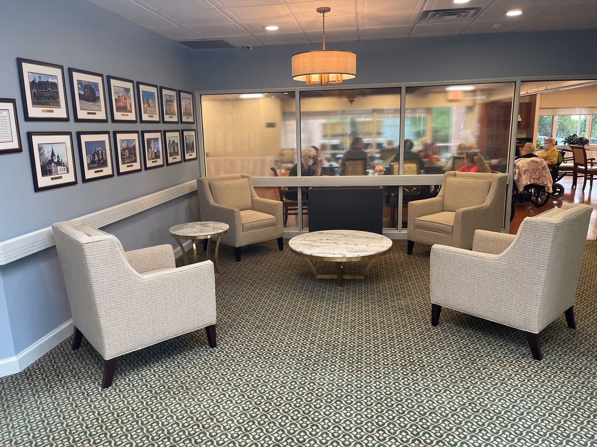 A seating area with four beige armchairs arranged around two round marble-top tables on a patterned carpet. The walls are painted light blue and decorated with framed pictures. Behind the seating area is a large glass window showing a dining room with several people seated at tables.