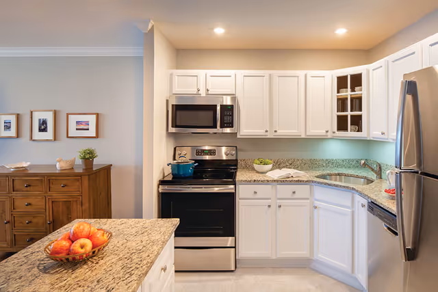 Modern kitchen with white cabinets, granite countertops, stainless steel appliances including a microwave, stove, and refrigerator. A bowl of apples is on the kitchen island, and a wooden sideboard with framed pictures and a small plant is visible in the adjacent room.