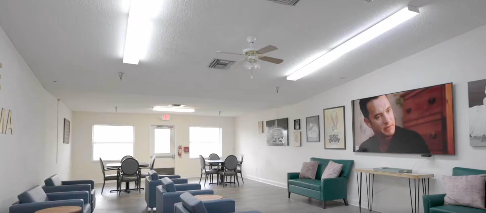 A bright communal living area with chairs and sofas arranged around small tables, a dining table in the back, wall art, and a wall-mounted TV.