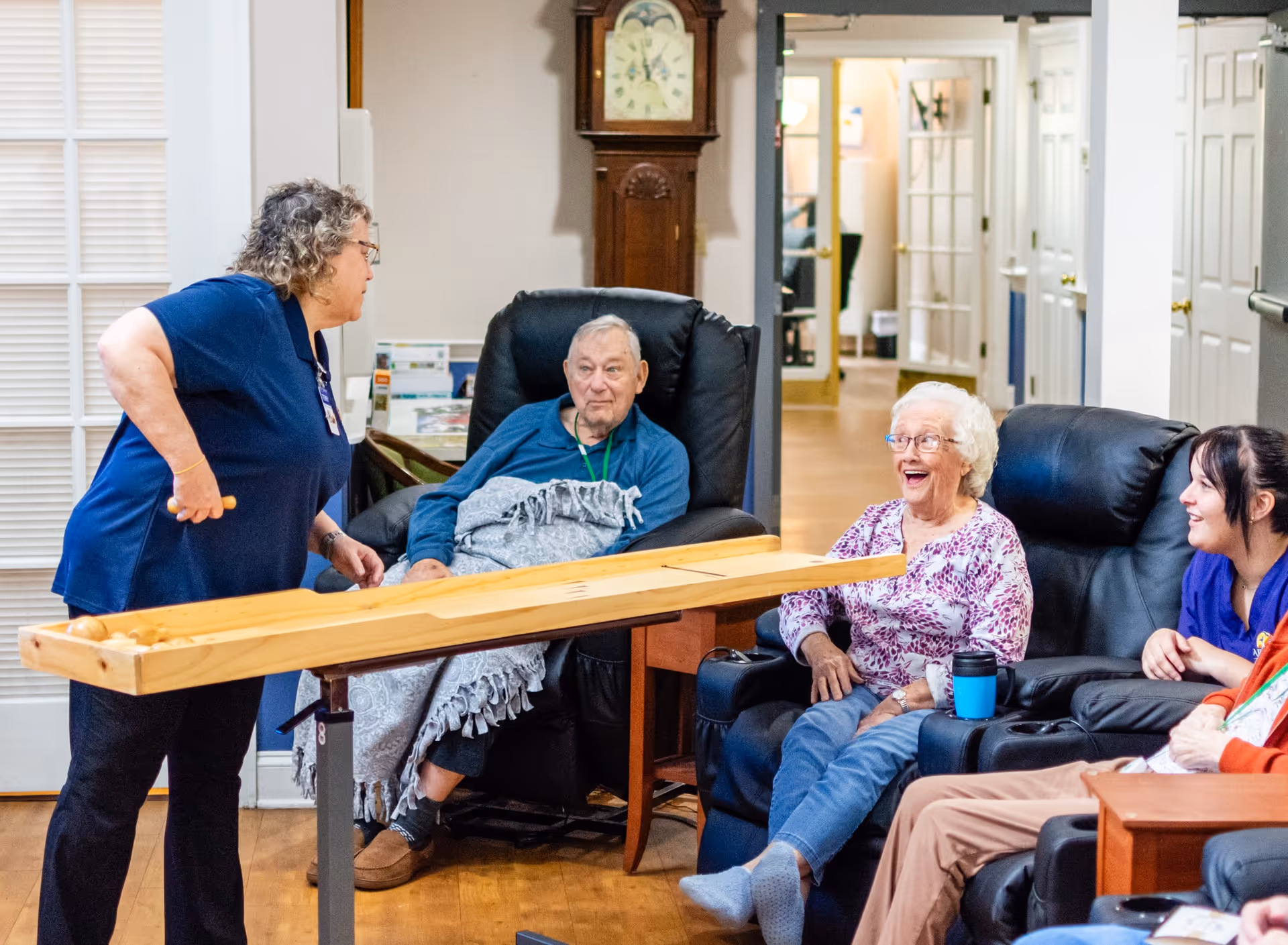 A staff member engages with elderly residents seated in recliners around a wooden activity table in a bright common lounge.