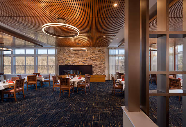 A spacious dining room with multiple tables covered in white tablecloths and set with red napkins and glassware. The room features large windows letting in natural light, a stone fireplace on the far wall, and modern circular ceiling lights. The ceiling has a wooden slat design, and the floor is carpeted in a dark pattern.