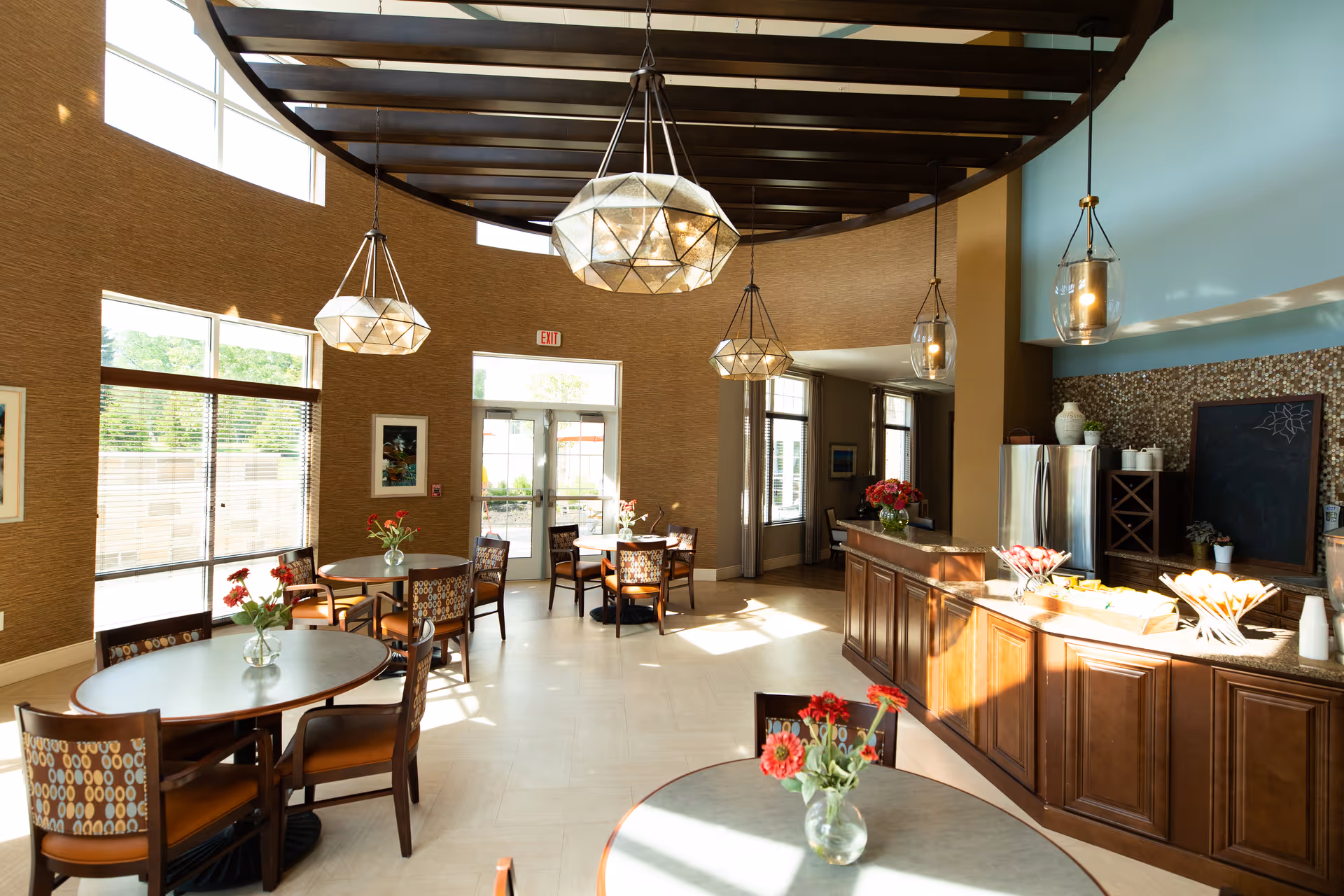 Sunlit communal dining area with round tables and chairs, pendant lights, floral centerpieces, and a wooden service counter.